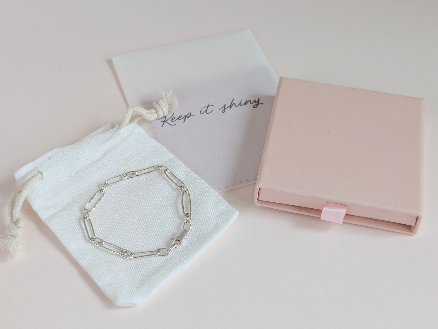 Silver bracelet on a white drawstring bag with 'Keep it shining' text, next to a pink jewelry box on a light pink background.