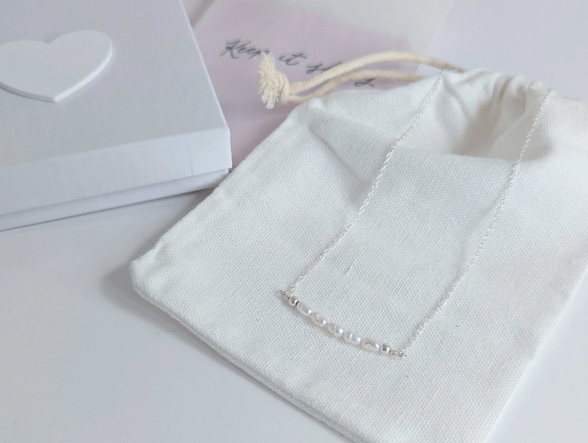 Dainty freshwater seed pearl bar necklace - shows packaging, including white cotton drawstring bag, white presentation box with white wooden heart on the lid and polishing cloths