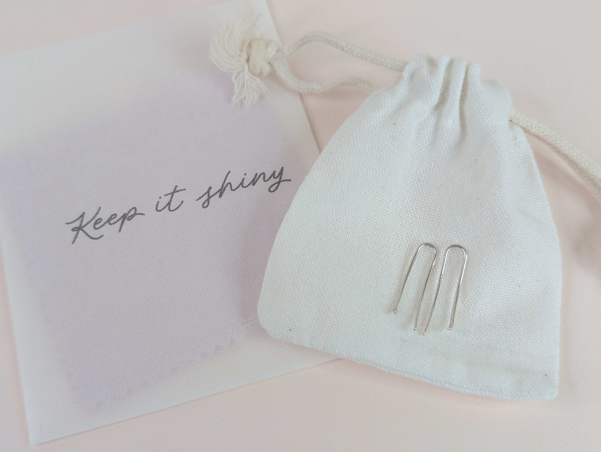White drawstring bag with silver ear climber earrings and a bag saying 'Keep it shiny' containing polishing cloths, on a light pink background