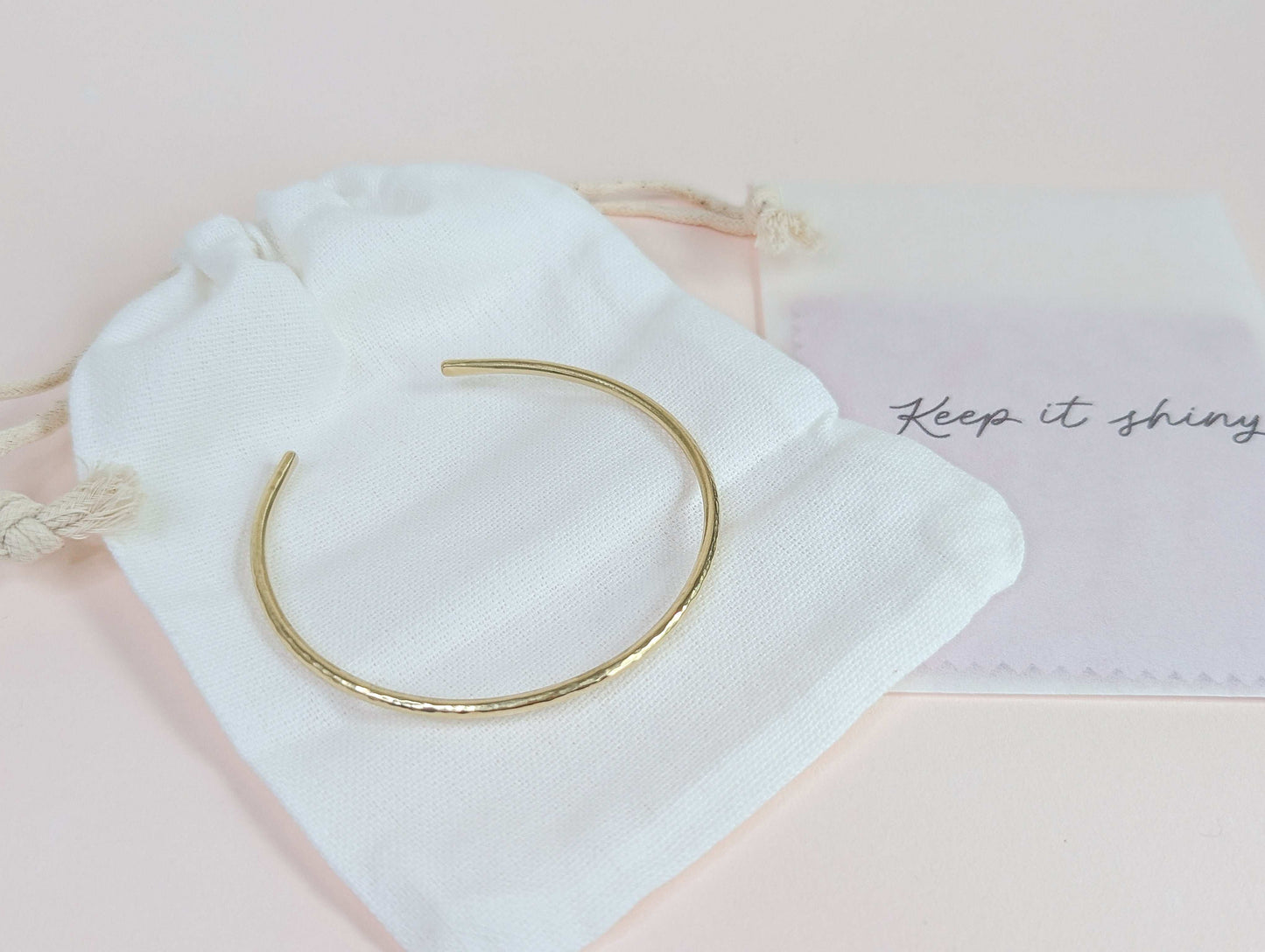 Gold bracelet on a white drawstring bag with 'Keep it shiny' polishing cloths on a light pink background