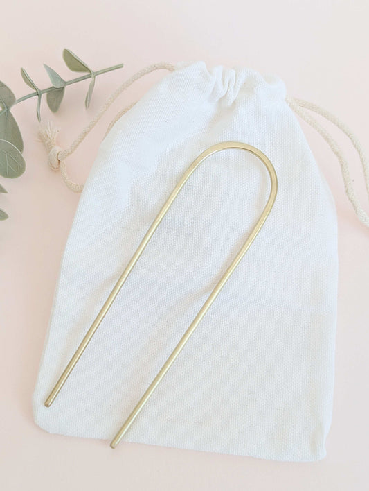Brushed brass gold round pin hair fork - laid flat on a white drawstring cotton bag