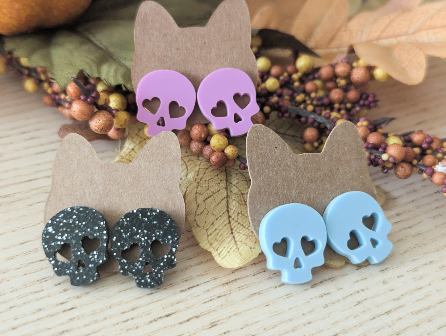 Skull-shaped earrings with heart cutouts on cardboard backing, surrounded by autumn leaves and berries.