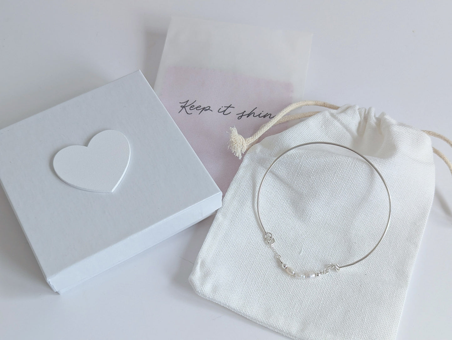 Thin sterling silver dainty cuff bangle with seed pearl bar chain - shows packaging including white drawstring cotton back, white jewellery presentation box with white wooden heart on the lid and polishing cloths