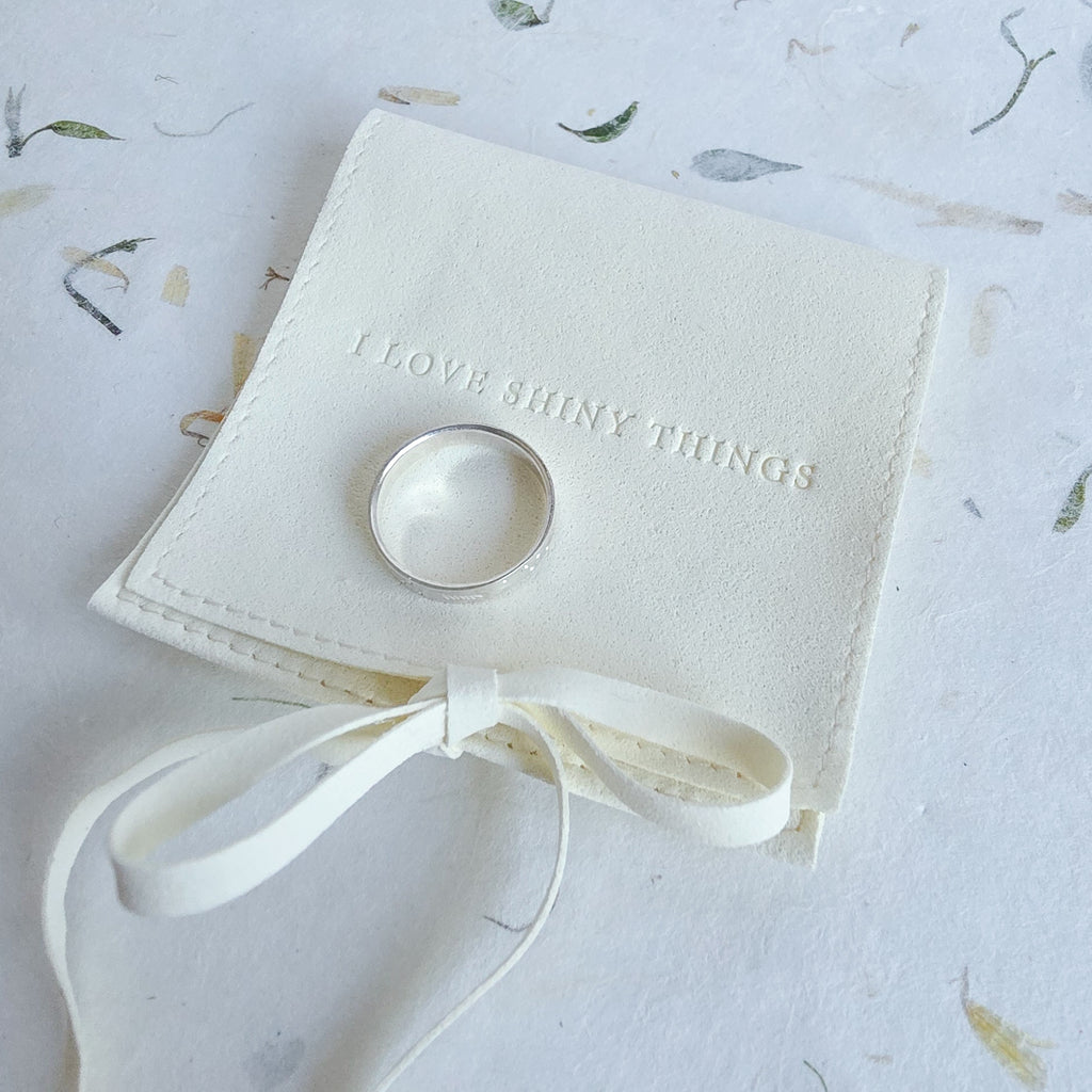A silver band ring displayed on a small cream-colored fabric ring box with a ribbon bow, placed on a light patterned surface. The box is embossed with the phrase 'I LOVE SHINY THINGS'.