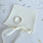 A silver band ring displayed on a small cream-colored fabric ring box with a ribbon bow, placed on a light patterned surface. The box is embossed with the phrase 'I LOVE SHINY THINGS'.