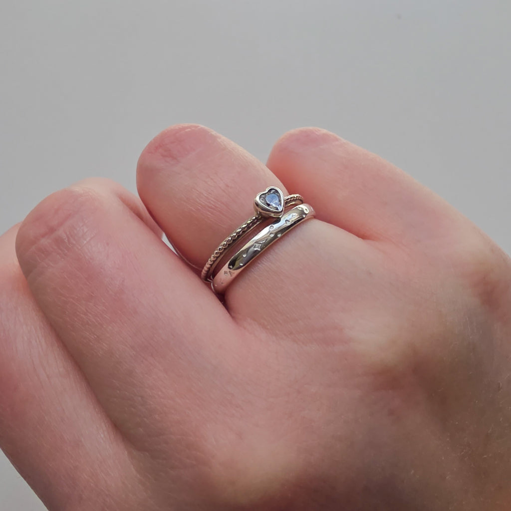 Hand wearing two silver rings with heart-shaped gemstones on a neutral background