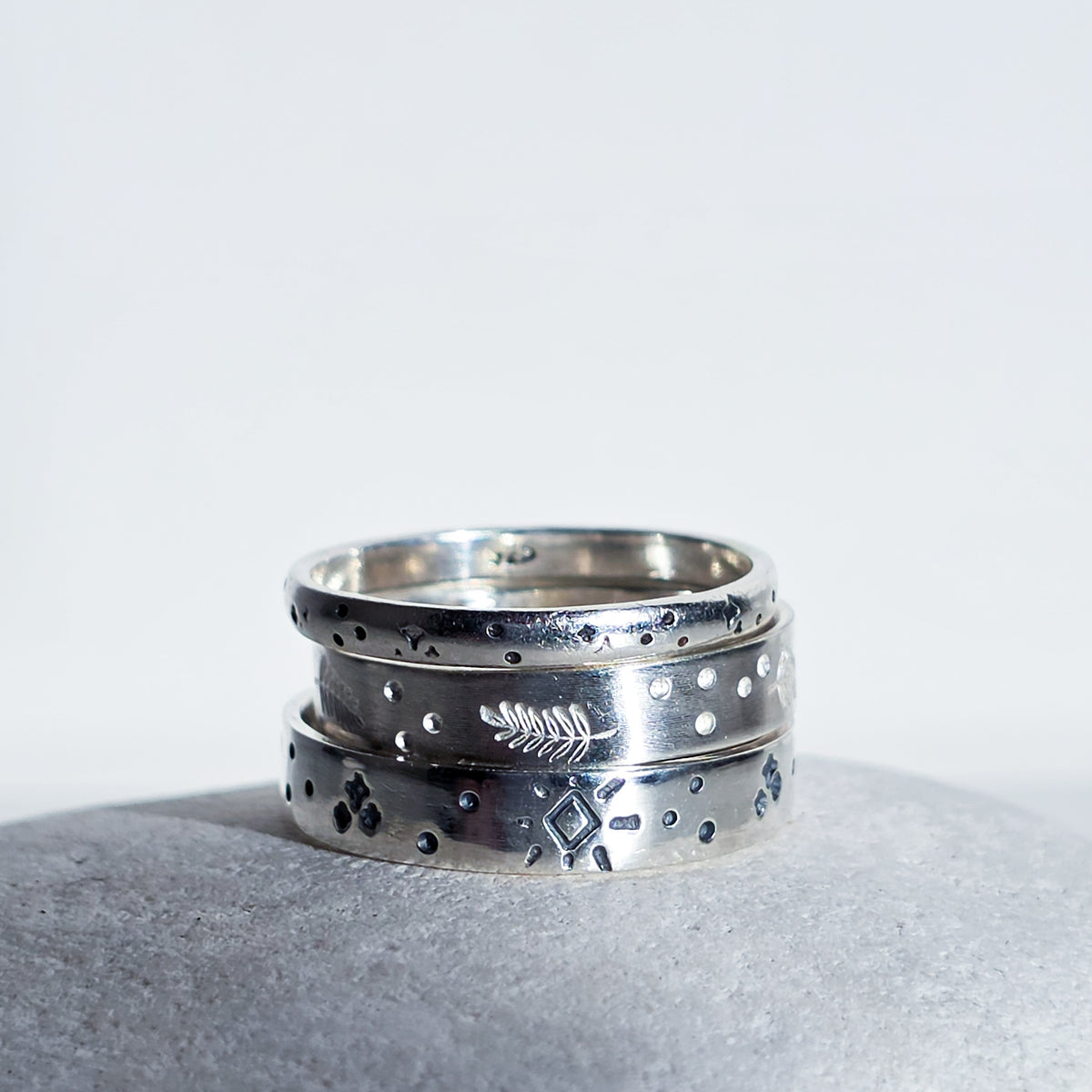 A stack of three silver rings with textured, hammered, and engraved details, resting on a gray stone against a white background.