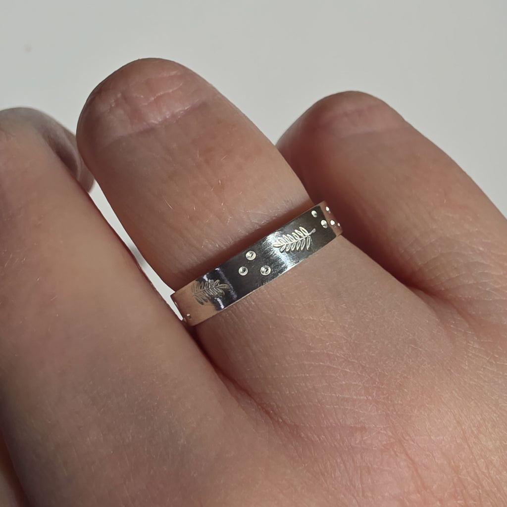 A silver band ring with engraved fern fronds and small raised dots, shown worn on a finger against a plain light background.
