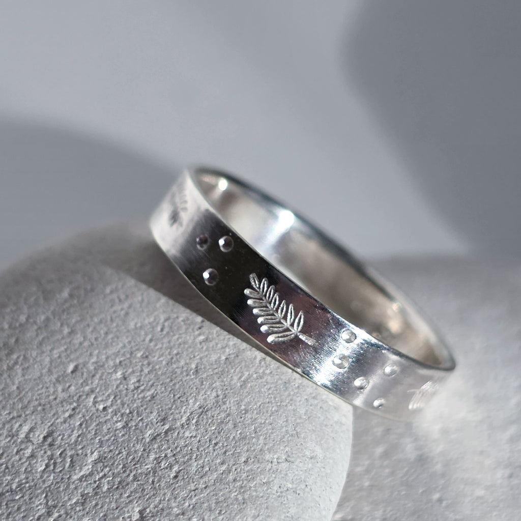 A sterling silver band ring with a textured, hammered finish, featuring a small engraved leaf motif and tiny raised dots, resting on a light gray stone surface.