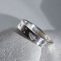 A sterling silver band ring with a textured, hammered finish, featuring a small engraved leaf motif and tiny raised dots, resting on a light gray stone surface.