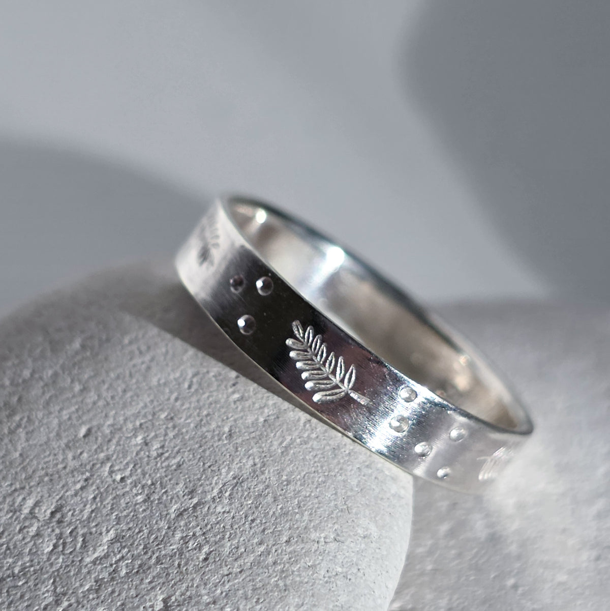 A sterling silver band ring with a textured, hammered finish, featuring a small engraved leaf motif and tiny raised dots, resting on a light gray stone surface.