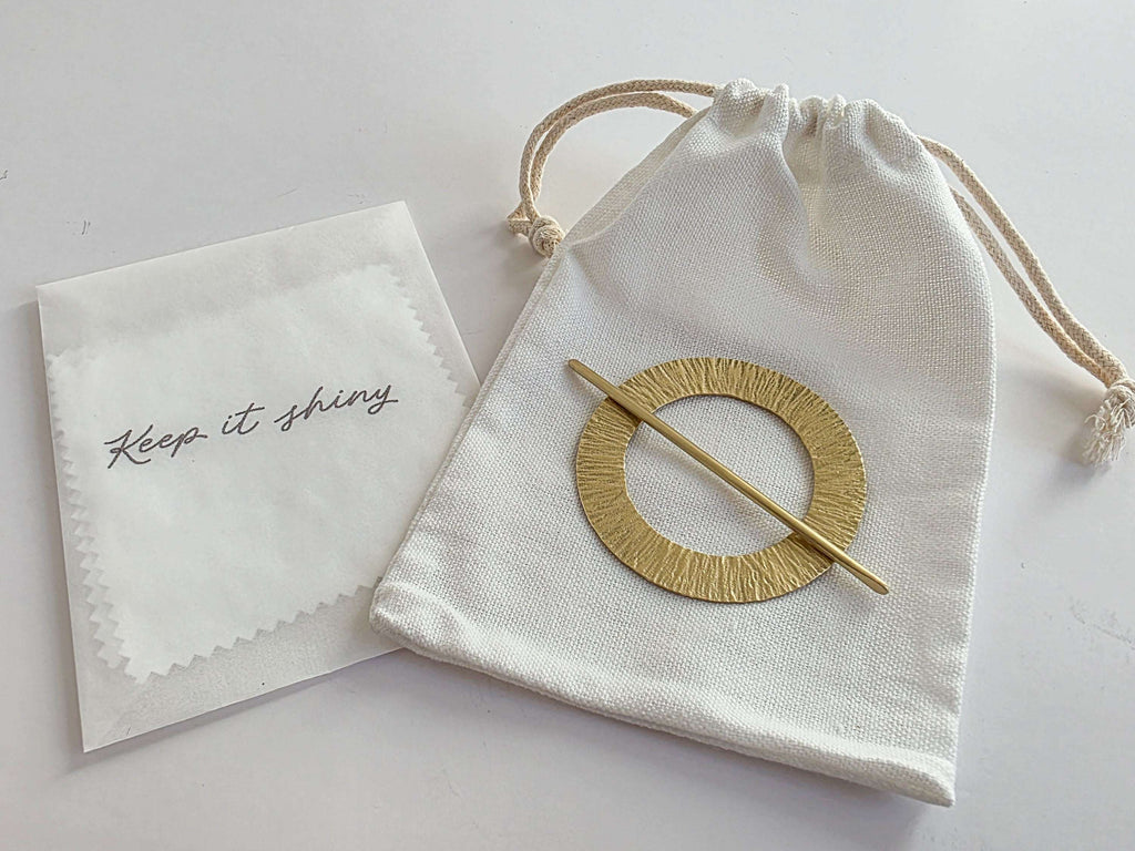 Gold circular brooch in a drawstring bag with a cloth labeled 'Keep it shiny' on a light background