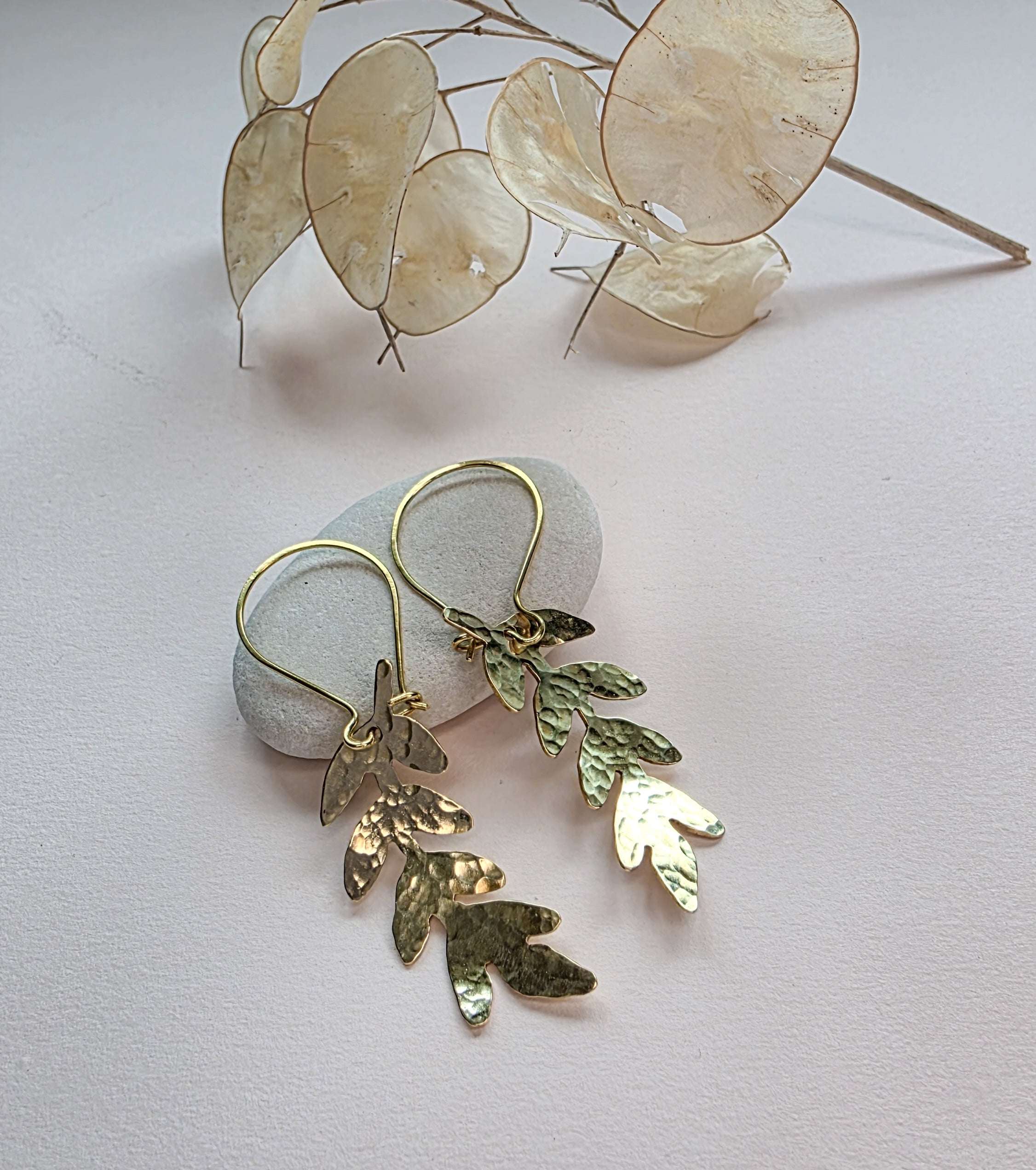 Gold leaf earrings on a light surface with dried leaves in the background