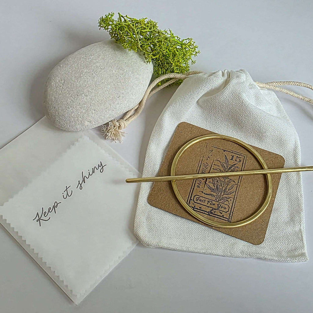 round brass hair barrette laid on a kraft card with a white cotton drawstring bag and polishing cloths