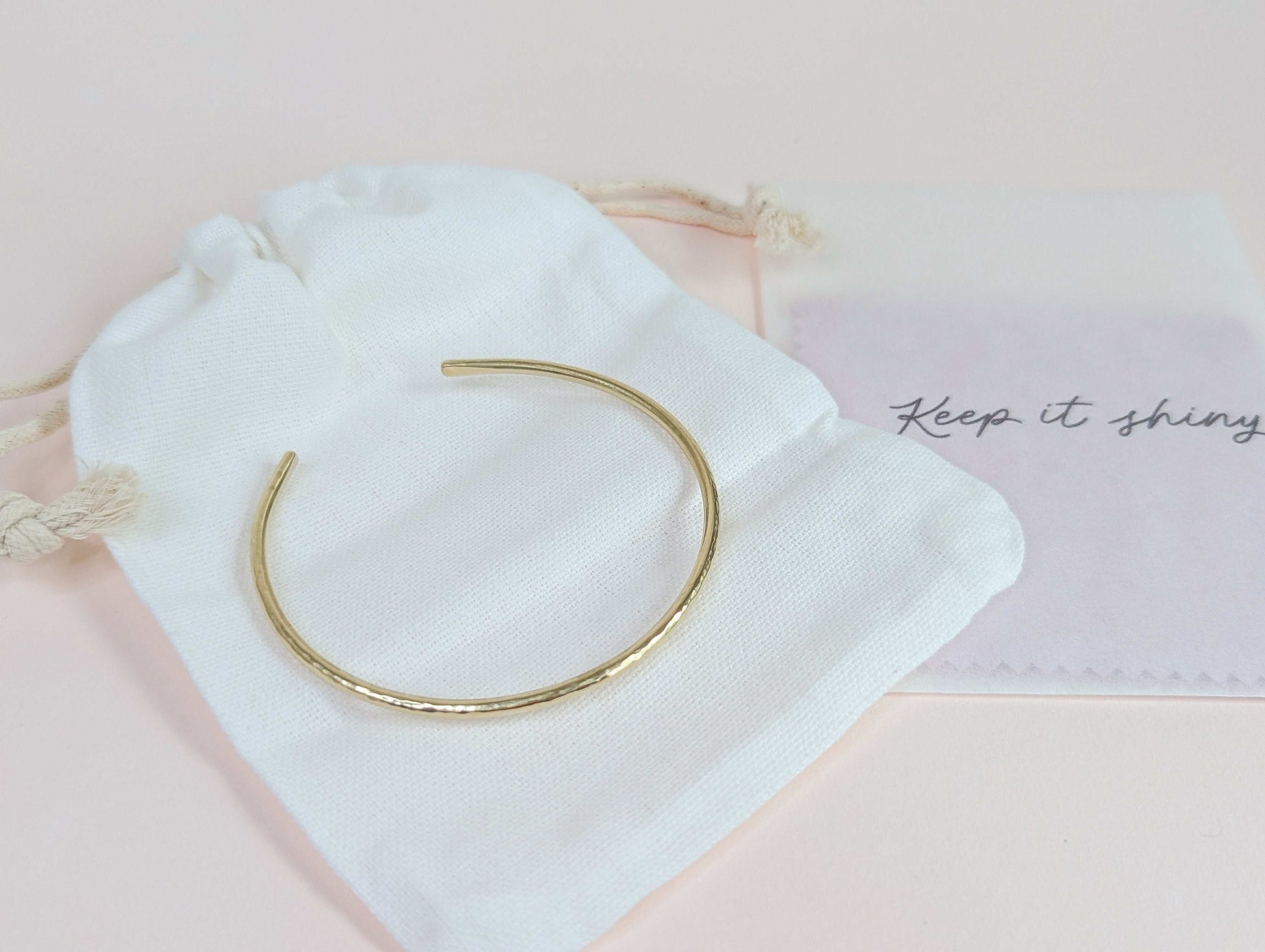 Gold bracelet on a white drawstring bag with 'Keep it shiny' polishing cloths on a light pink background