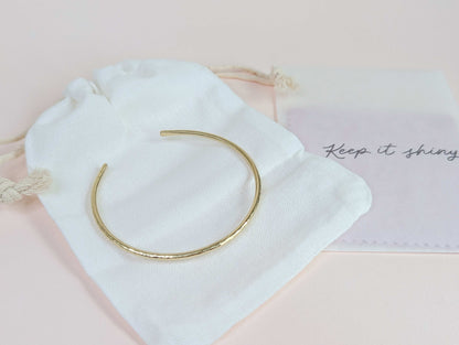 Gold bracelet on a white drawstring bag with 'Keep it shiny' polishing cloths on a light pink background