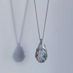Dendritic snow agate leaf Necklace with a teardrop-shaped pendant on a light grey surface on a chain against a light grey background