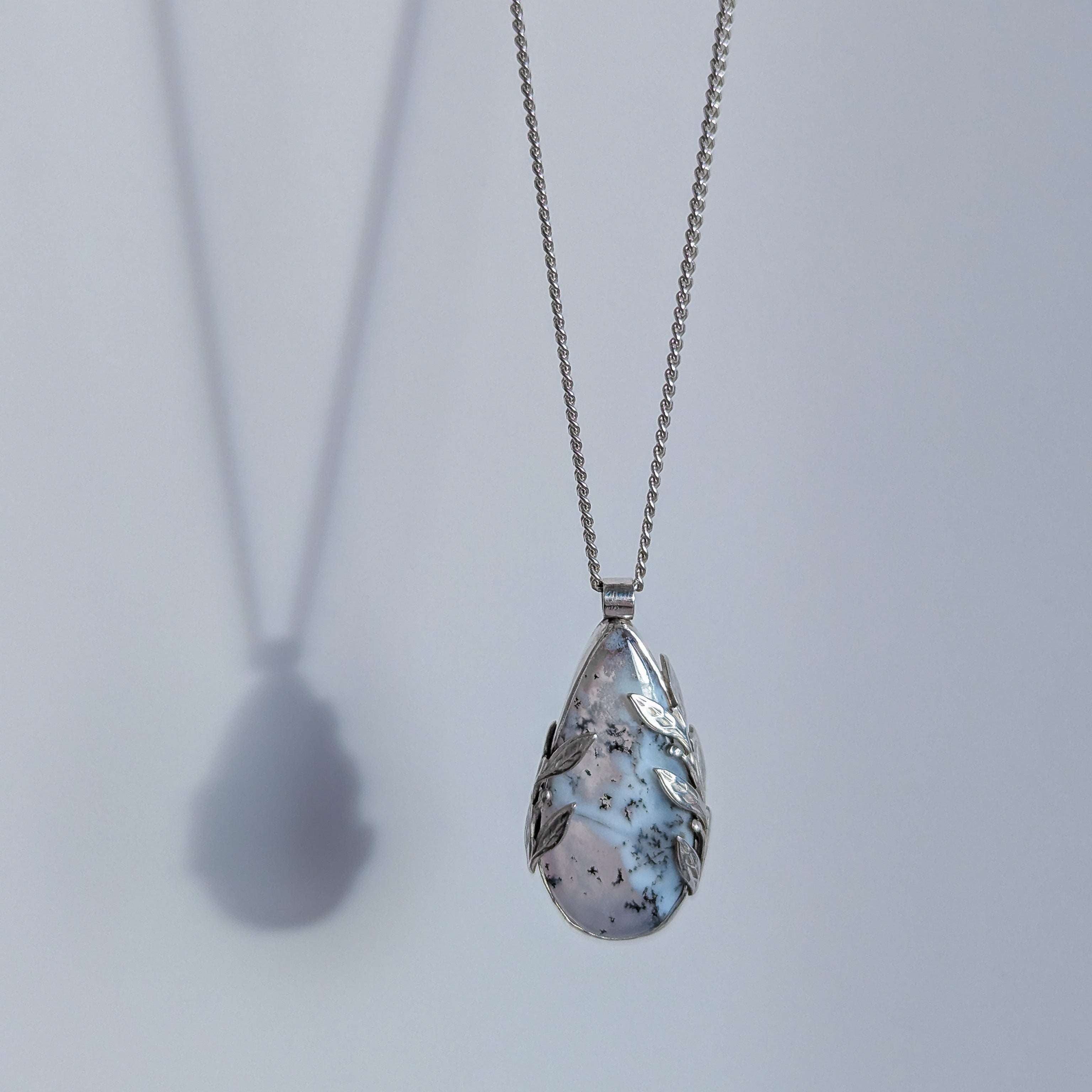 Dendritic snow agate leaf Necklace with a teardrop-shaped pendant on a light grey surface on a chain against a light grey background