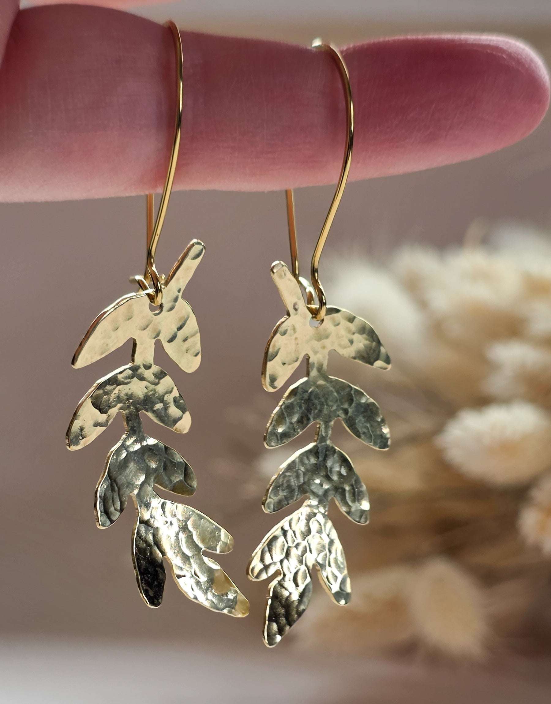 Gold leaf earrings held by a hand against a blurred natural background