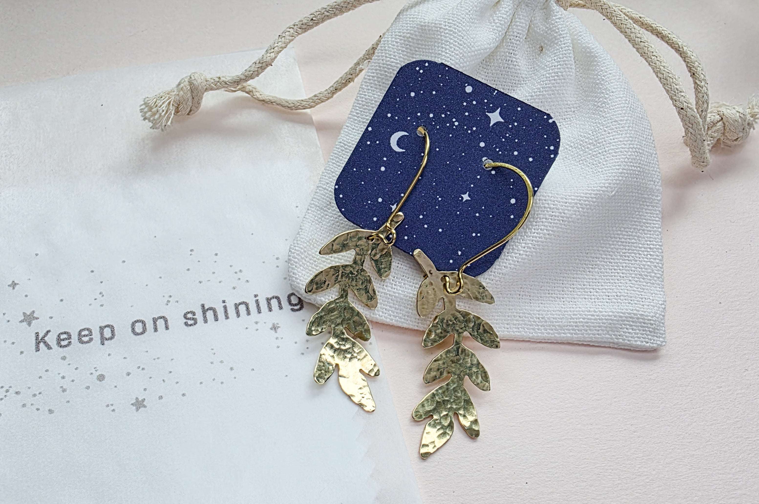 Gold leaf earrings on a white drawstring bag with a blue card and 'Keep on shining' text.