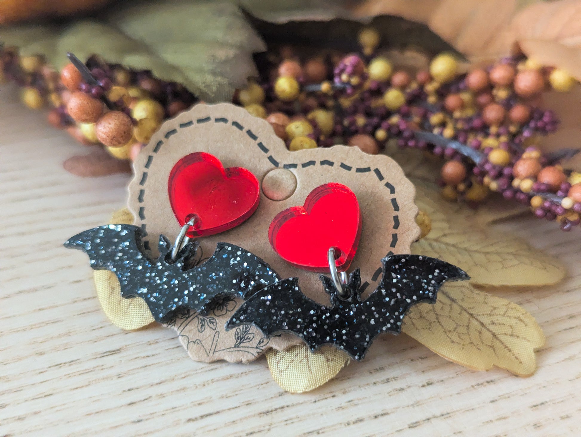 Pair of earrings with red heart and bat design on a wooden surface with decorative elements.
