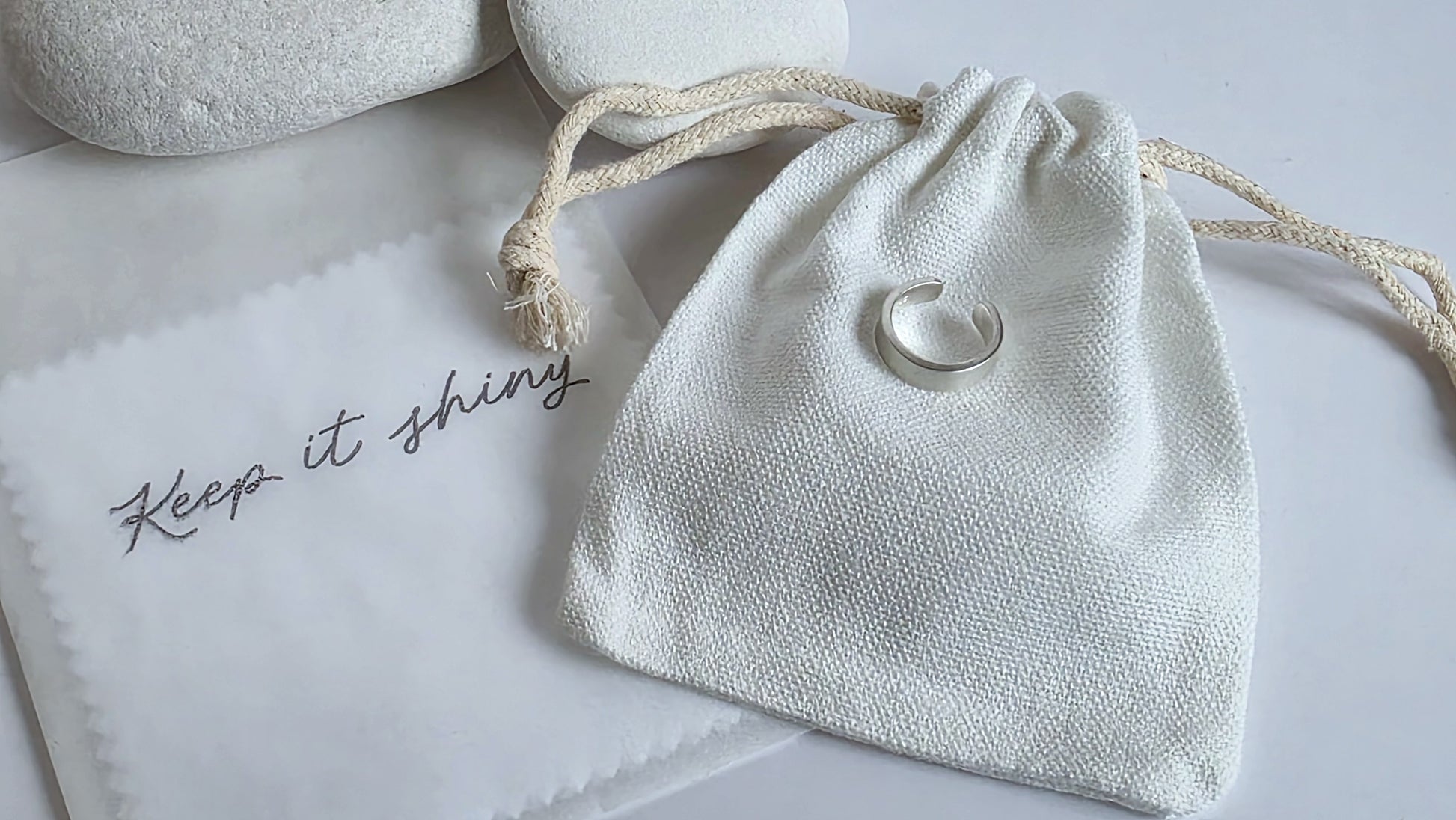 White drawstring bag with silver ear cuff on a light gray surface with 'Keep it shiny' text.