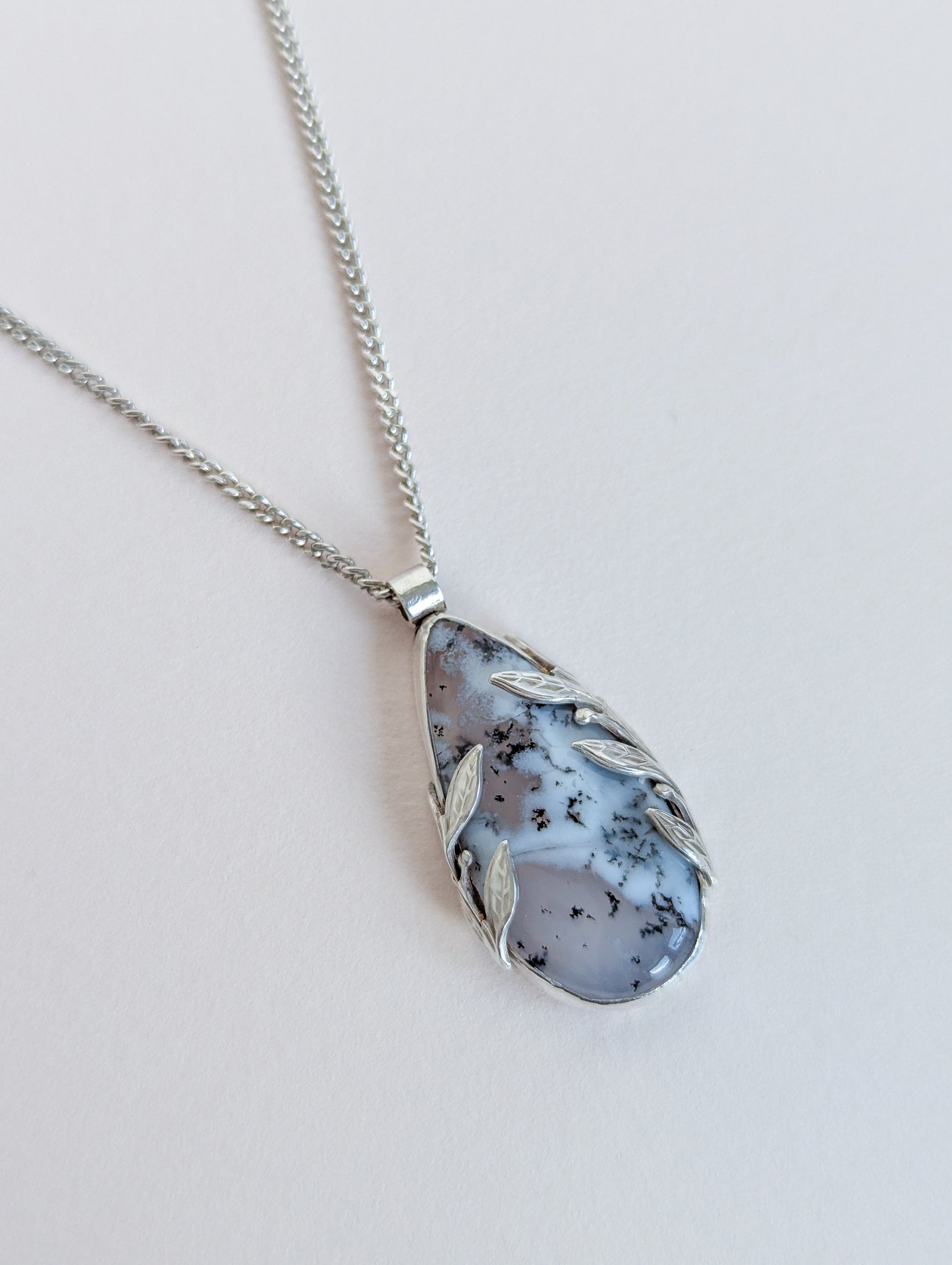 Teardrop-shaped pendant with leaf designs on a silver chain against a light background