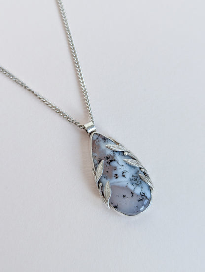 Teardrop-shaped pendant with leaf designs on a silver chain against a light background