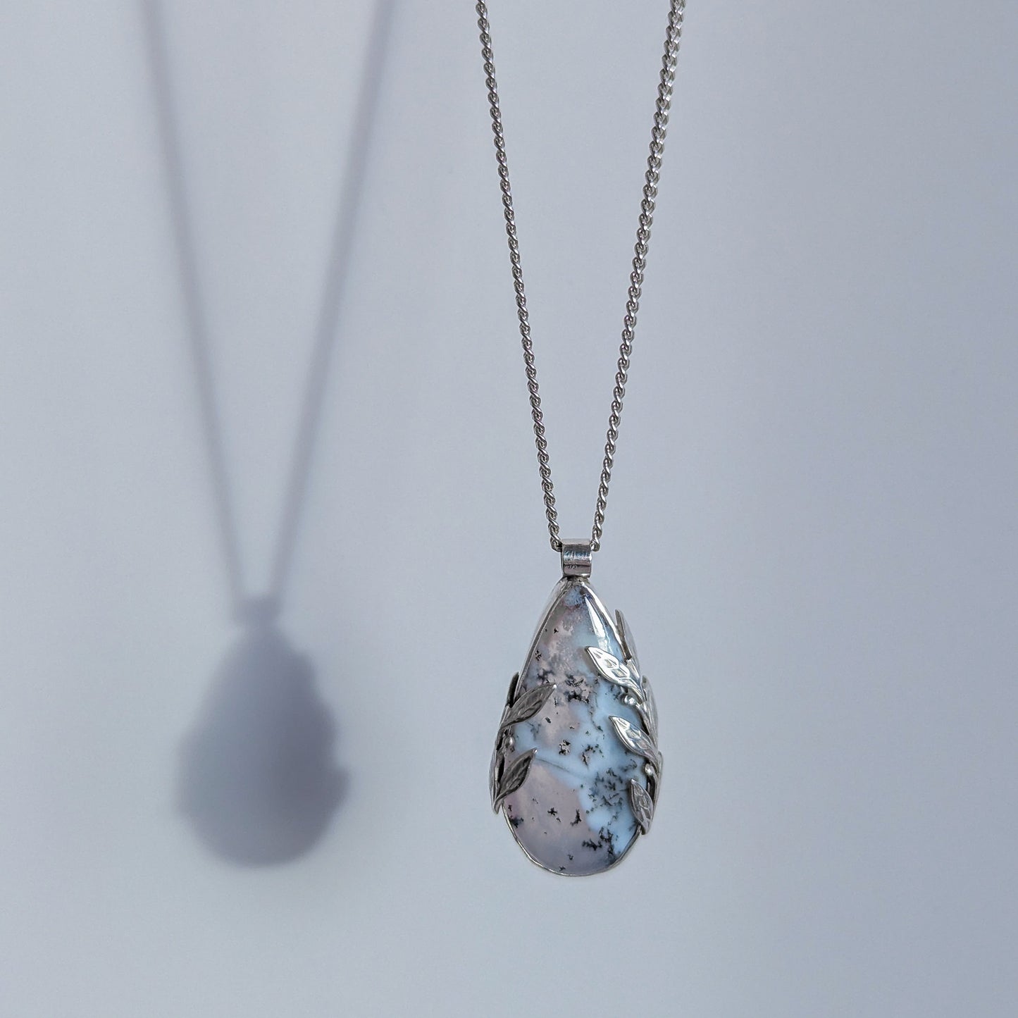 Dendritic snow agate leaf Necklace with a teardrop-shaped pendant on a light grey surface on a chain against a light grey background