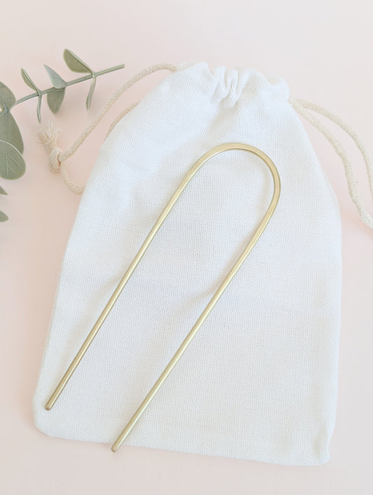 Brushed brass gold round pin hair fork - laid flat on a white drawstring cotton bag