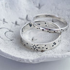 Two silver rings with hand-stamped black geometric and dotted patterns, resting on a textured light gray stone dish.