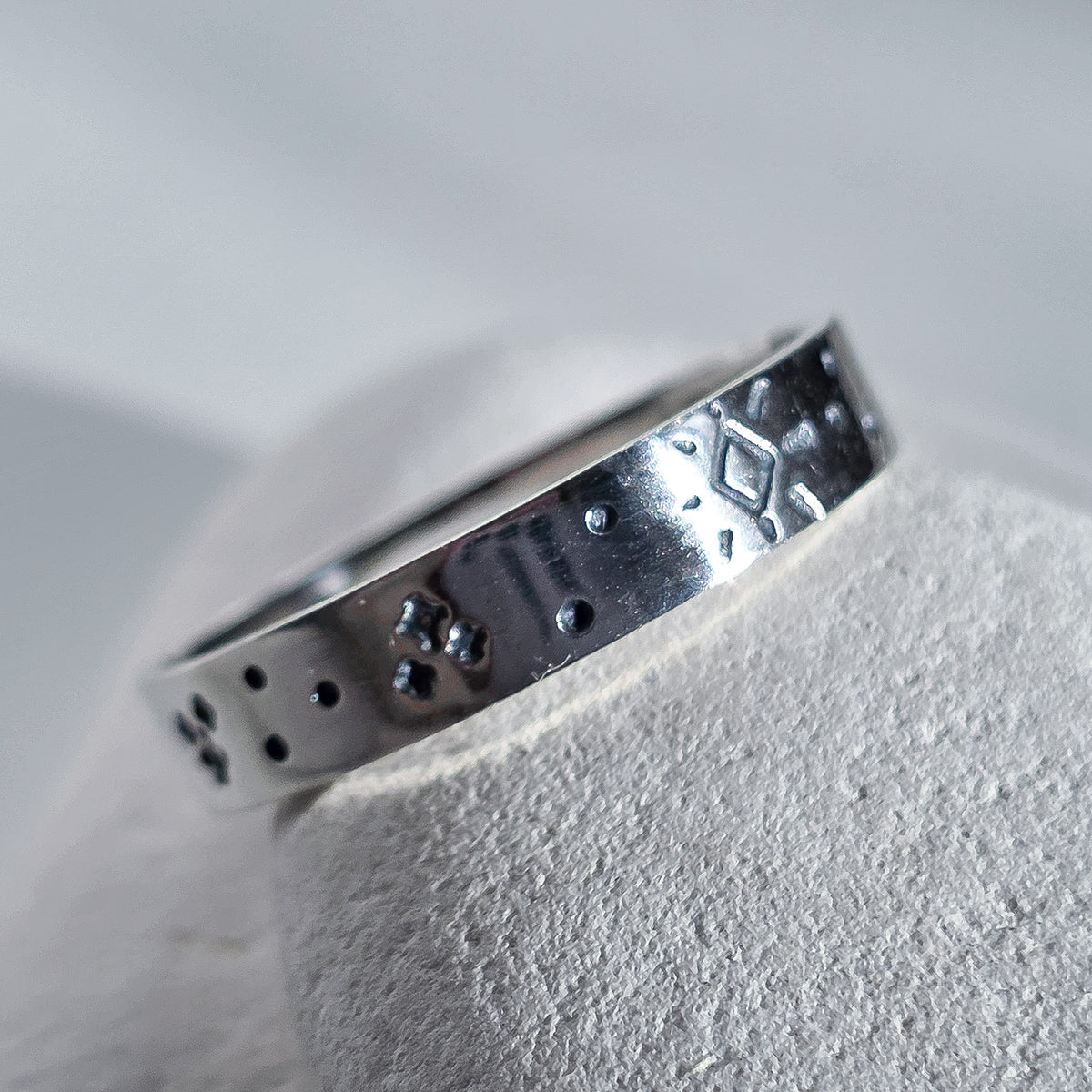 A sterling silver band ring with textured geometric and dotted patterns, resting on a textured light gray display stand.