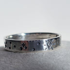 A sterling silver band ring with hand-stamped geometric and dotted patterns, resting on a textured gray surface against a light gray background.