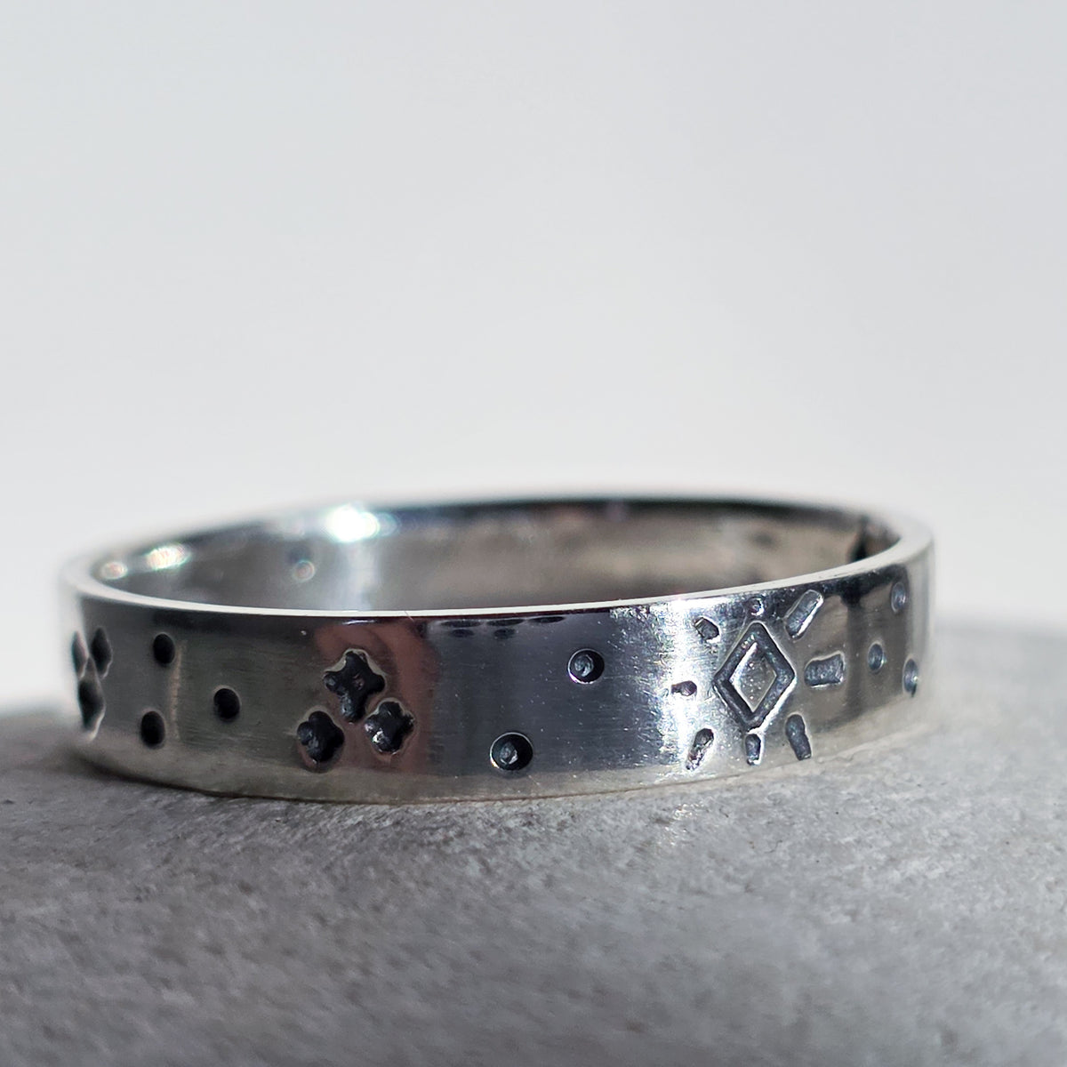 A sterling silver band ring with hand-stamped geometric and dotted patterns, resting on a textured gray surface against a light gray background.