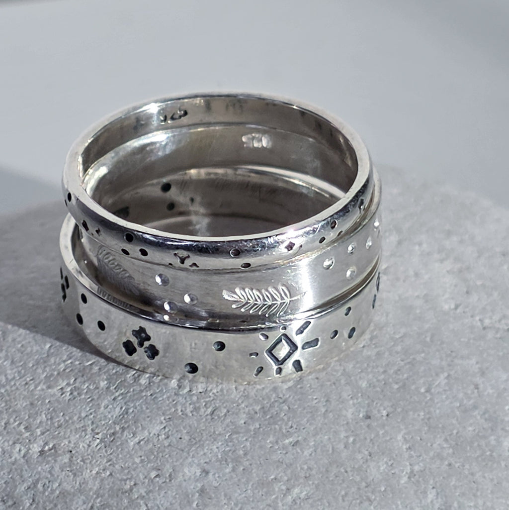 A pair of stacked sterling silver rings with engraved geometric and natural motifs, resting on a textured gray stone surface.