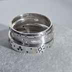 A pair of stacked sterling silver rings with engraved geometric and natural motifs, resting on a textured gray stone surface.