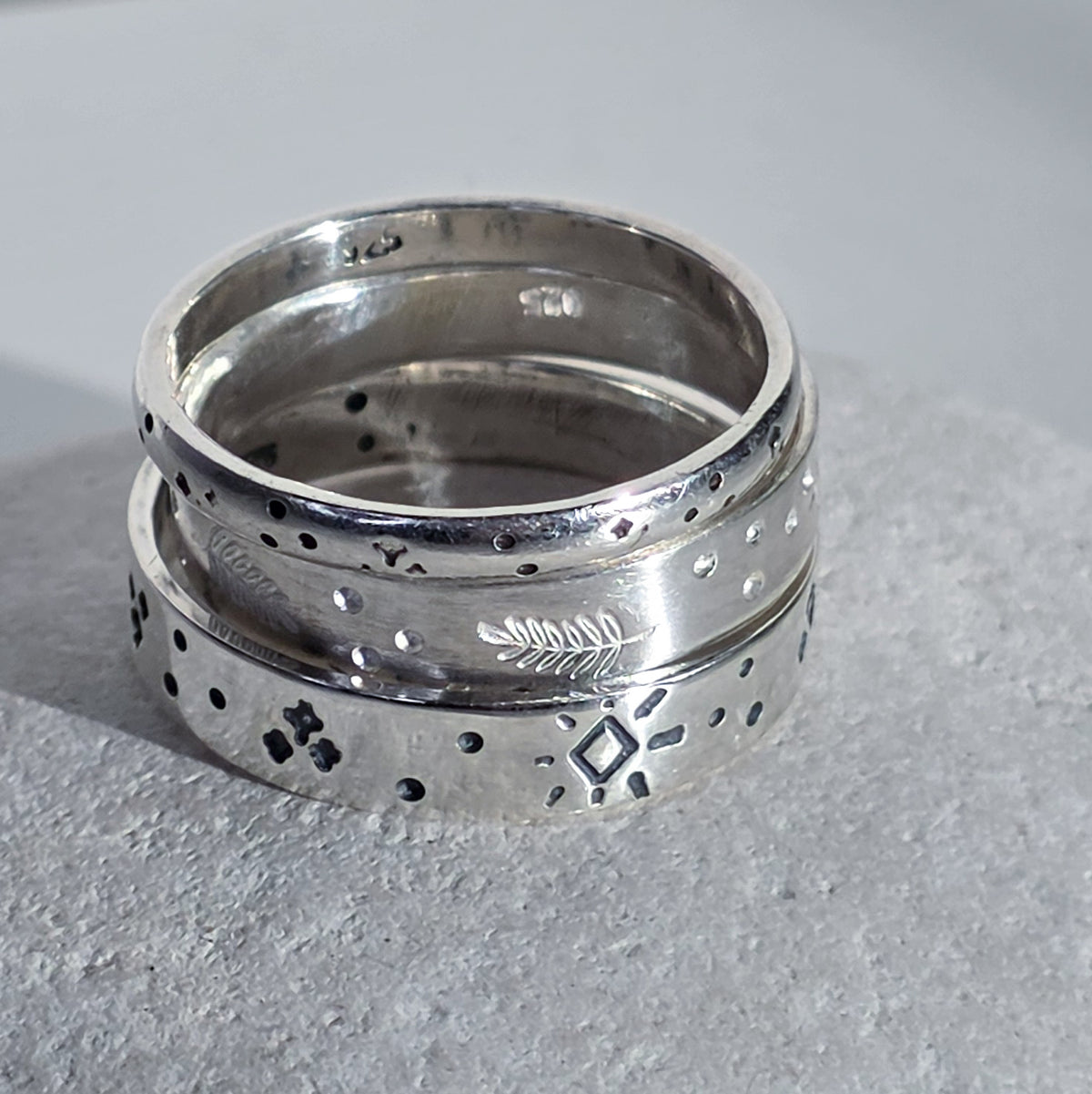 A pair of stacked sterling silver rings with engraved geometric and natural motifs, resting on a textured gray stone surface.