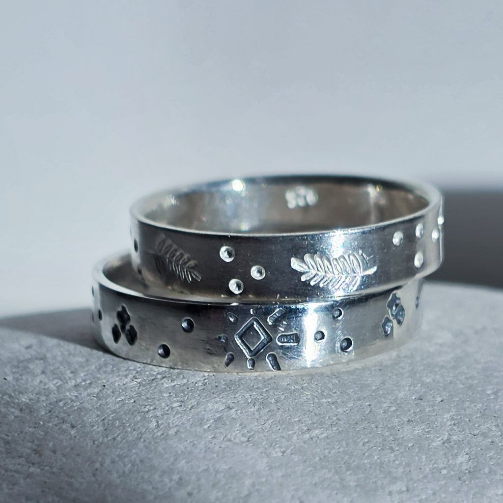 Two stacked sterling silver rings with engraved geometric and leaf-like patterns, displayed on a textured gray surface against a neutral gray background.