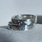 Two stacked sterling silver rings with engraved geometric and leaf-like patterns, displayed on a textured gray surface against a neutral gray background.